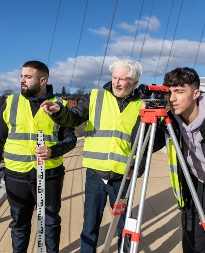 Three individuals wearing high-visibility vests conducting a land surveying exercise on a bridge. One is looking through a theodolite, while another holds a measuring staff, and a lecturer observes under a bright blue sky. Three individuals wearing high-visibility vests conducting a land surveying exercise on a bridge. One is looking through a theodolite, while another holds a measuring staff, and a lecturer observes under a bright blue sky.